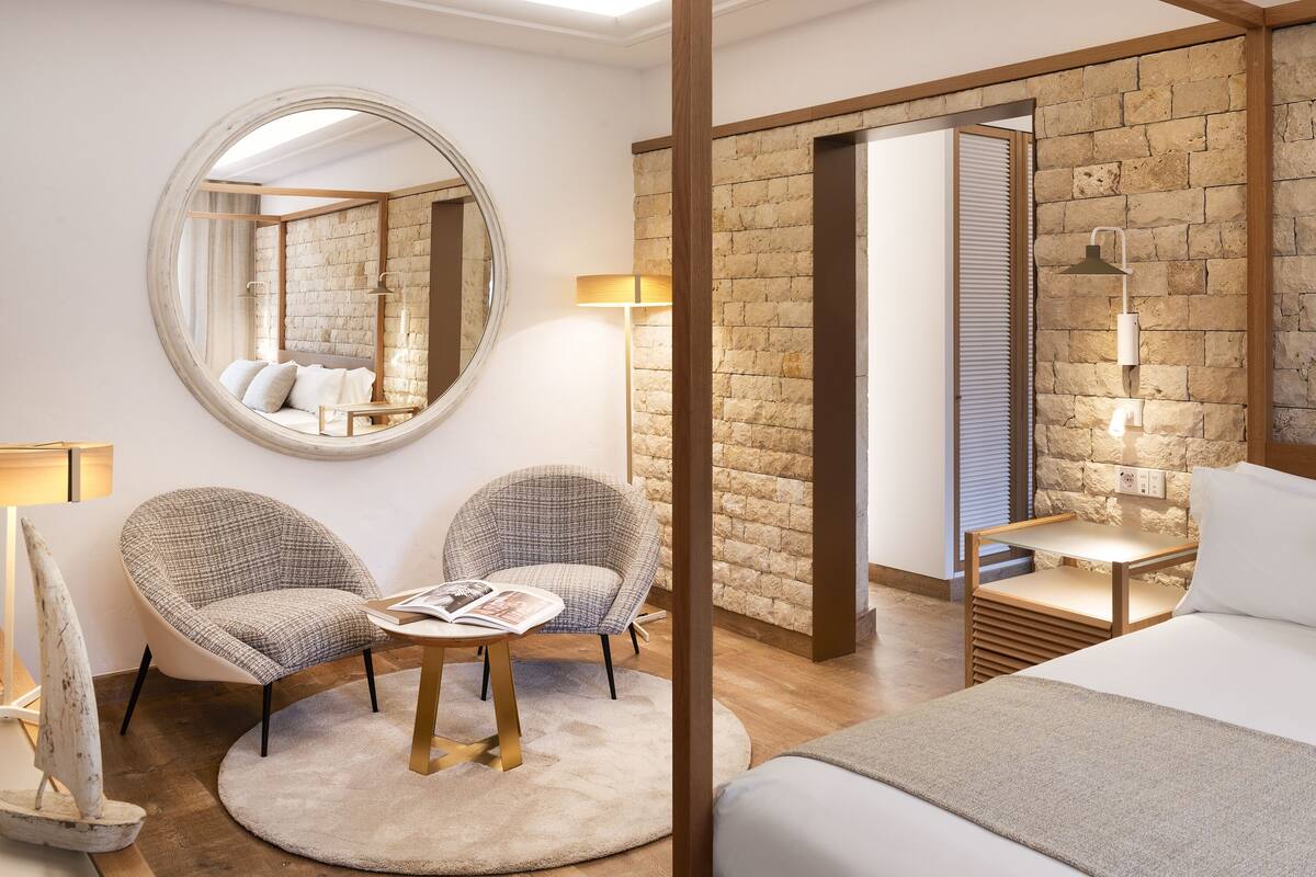 Une chambre d’hôtel moderne avec un grand miroir rond, deux fauteuils, une petite table avec un magazine, un lit doté d’une tête de lit en bois et un mur en pierre.