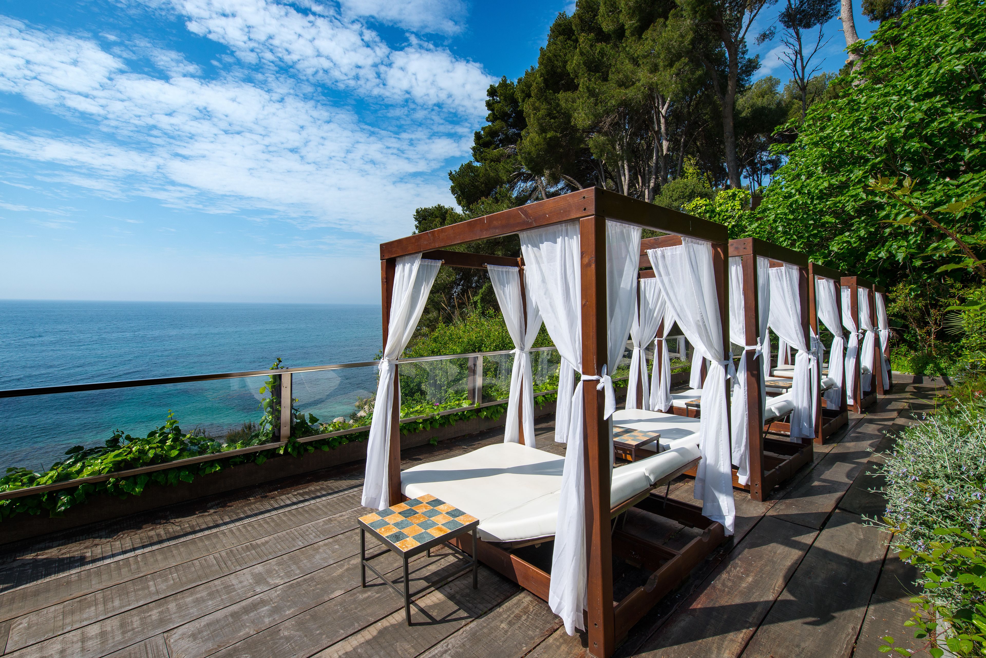 A row of wooden canopy beds with white curtains, a small table, and a view of the sea.