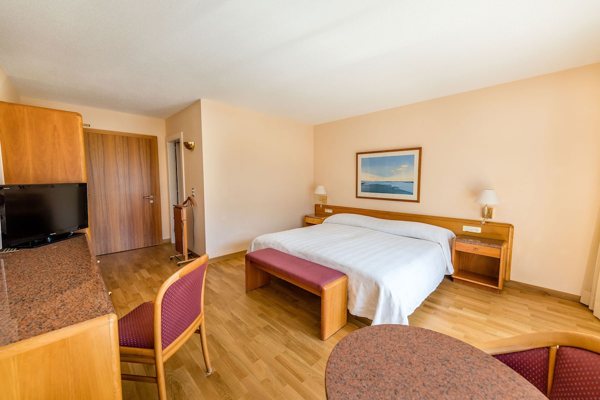 A hotel room with a bed, a desk, a TV, a bench, and a painting on the wall.