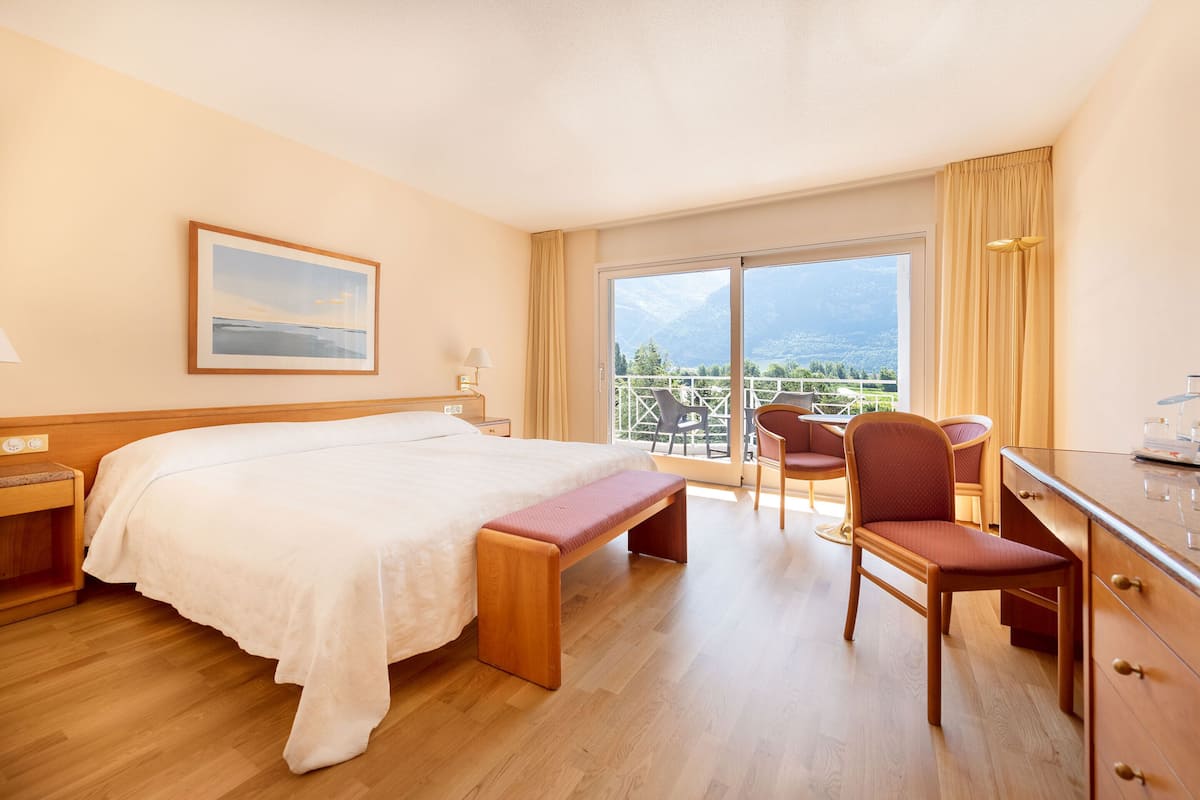 A hotel room with a bed, a desk, a chair, and a balcony with a view of mountains.