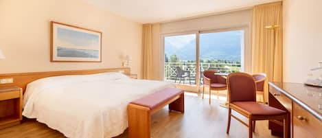 A hotel room with a bed, a desk, a chair, a bench, a window with a view of mountains, and a painting on the wall.