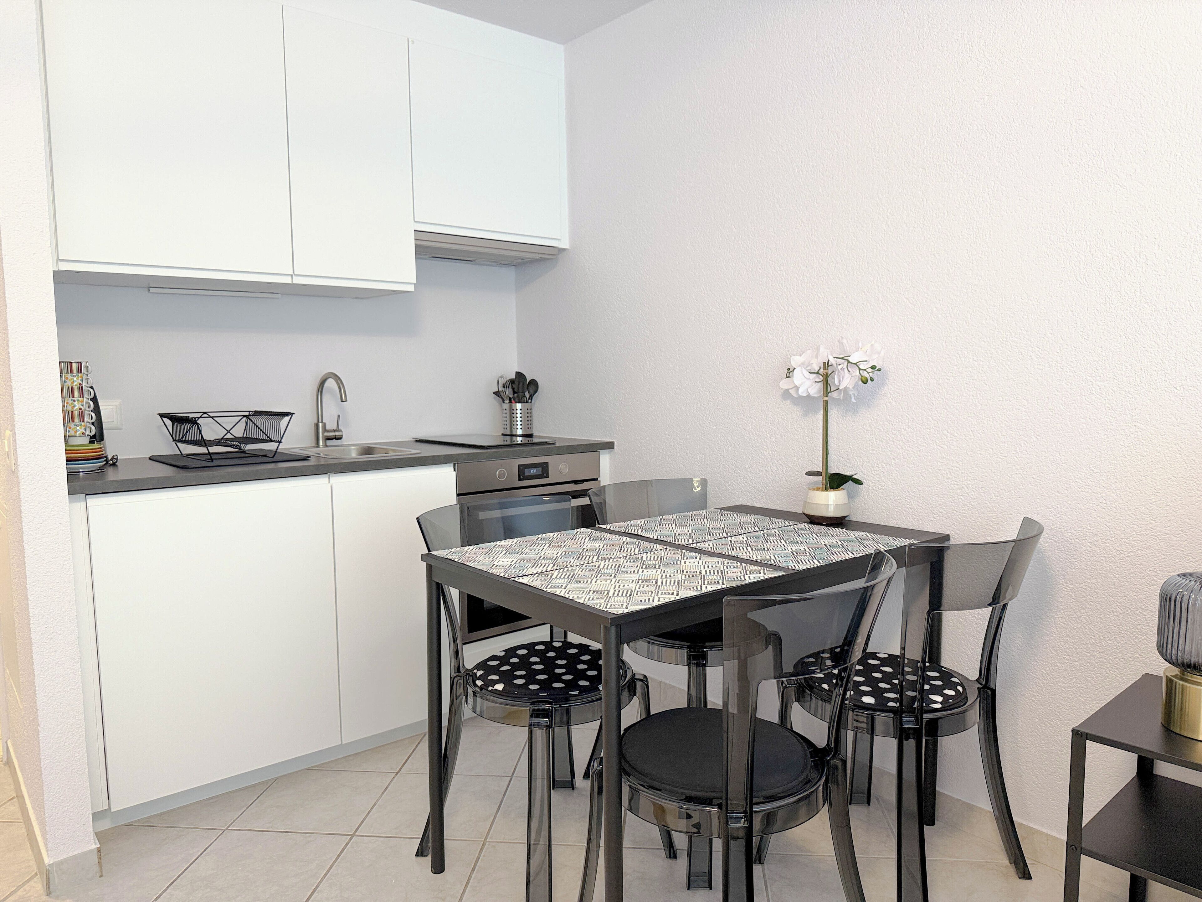 Een kleine keuken met witte kasten, een zwart aanrecht en een eethoek met een tafel met dessin en zwarte stoelen.