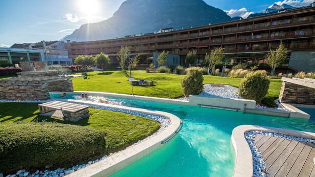 Et hotel med et poolområde, omgivet af velplejede græsplæner og med et bjergrigt landskab i baggrunden.