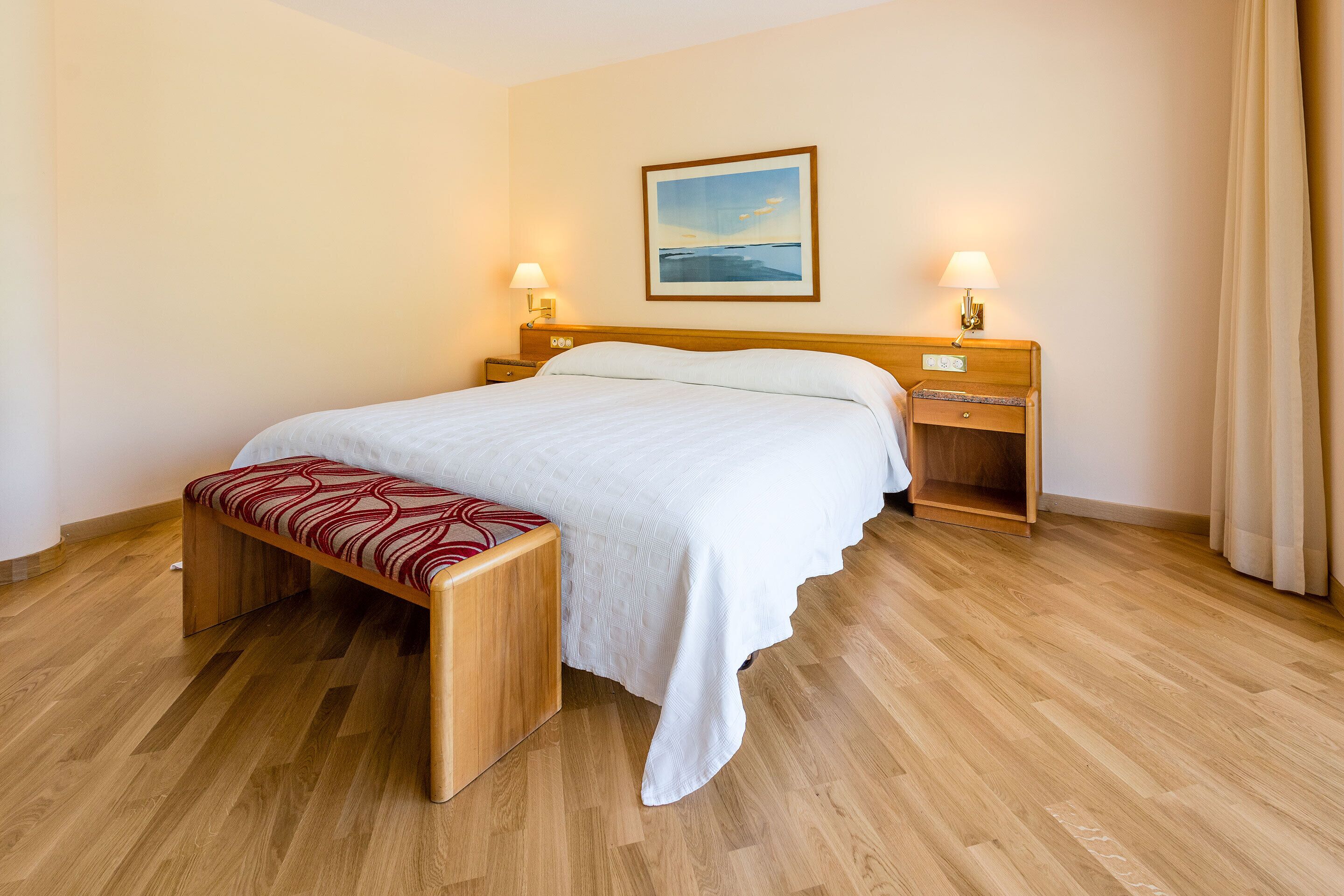 Habitación de hotel con cama de madera, un banco, dos mesitas de noche con lámparas y un cuadro enmarcado en la pared.