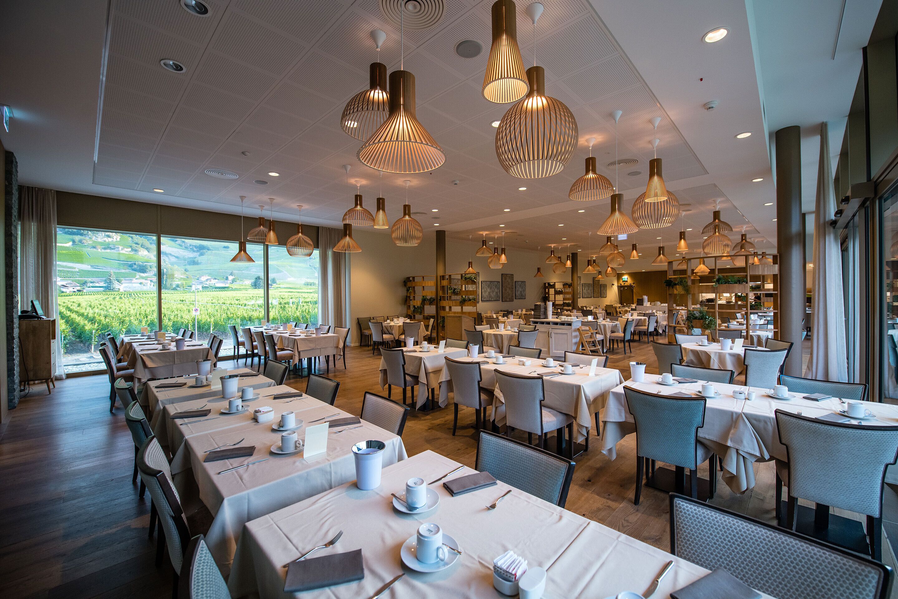 Un vaste espace dédié à la salle à manger, avec de nombreuses tables dressées, de grandes suspensions décoratives et une vue sur un vignoble grâce à de larges fenêtres.