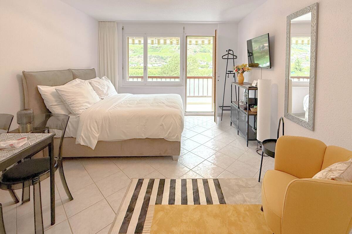 A bedroom with a bed, a desk, a chair, a TV, and a balcony with a view of greenery.