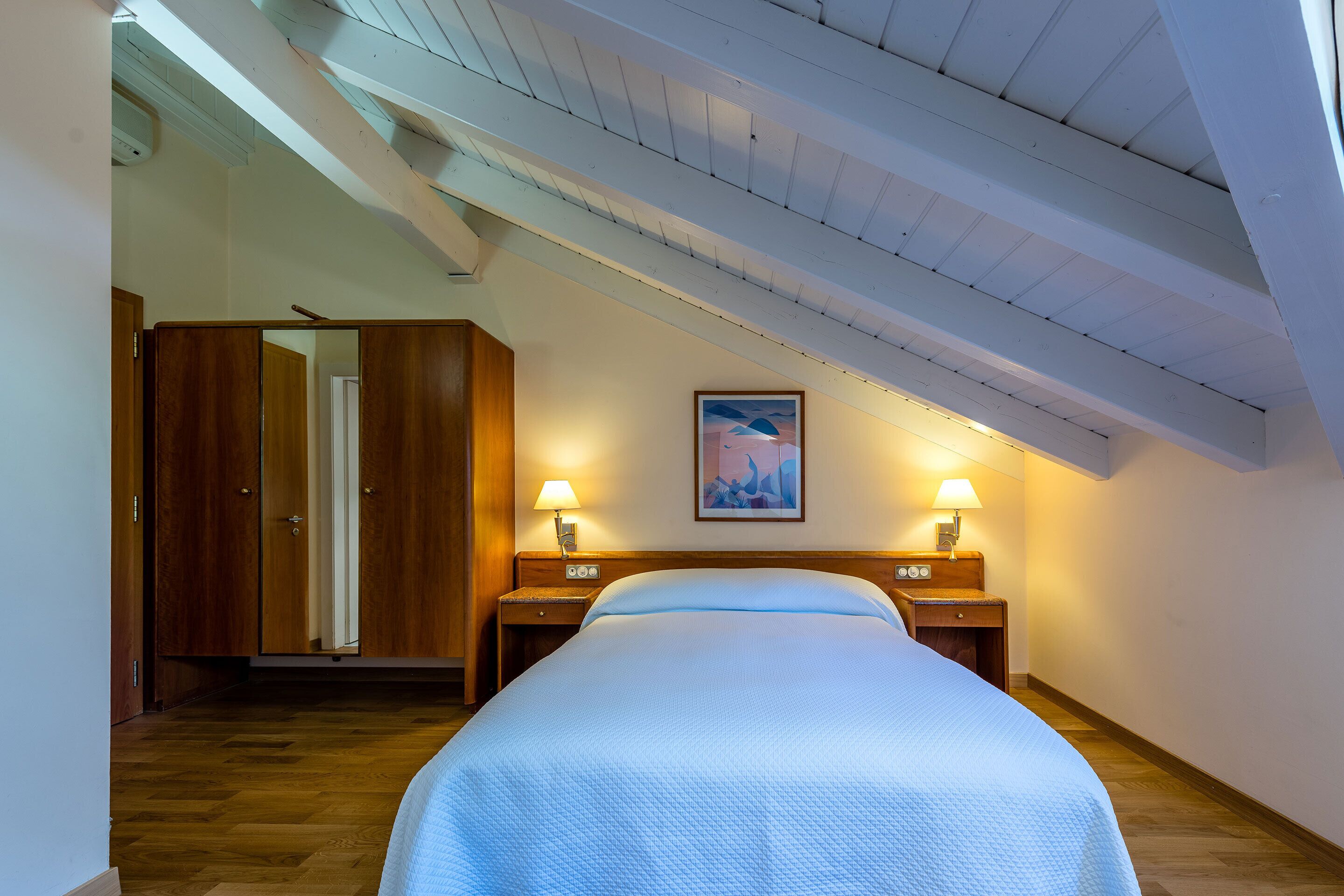 A bedroom with a large bed, wooden wardrobe, two bedside tables with lamps, and a ceiling with exposed beams.