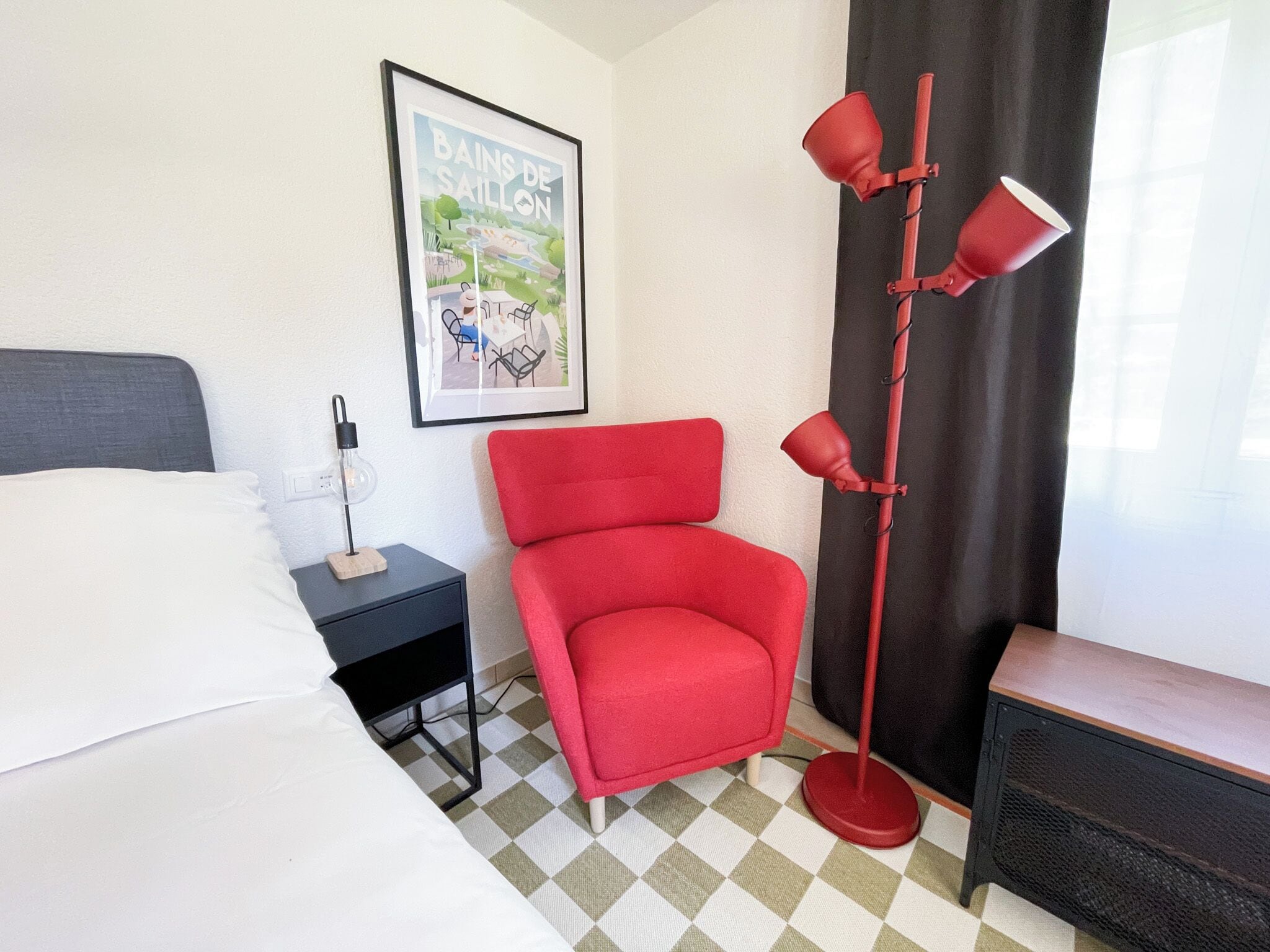 A hotel room with a bed, a red chair, a nightstand with a lamp, a framed picture on the wall, and a checkered floor.