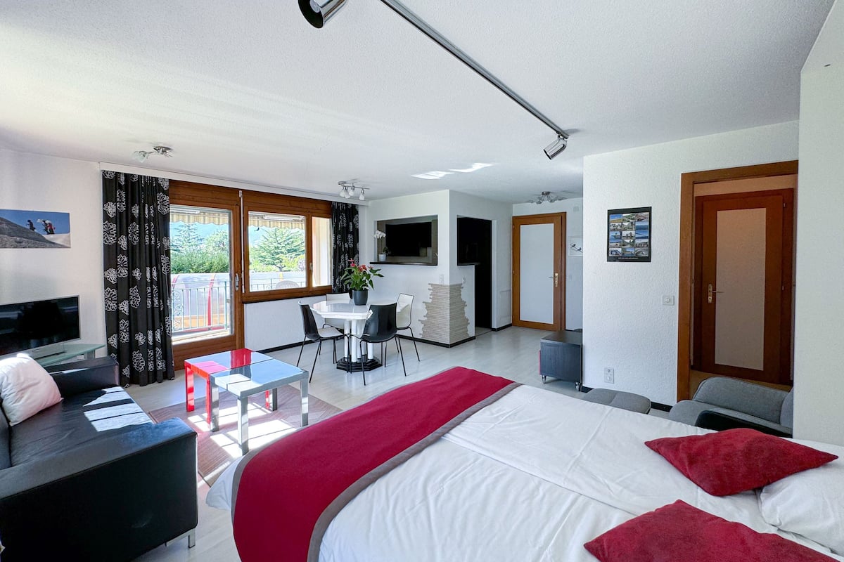 A modern hotel room with a bed, a sofa, a dining table, and a balcony.