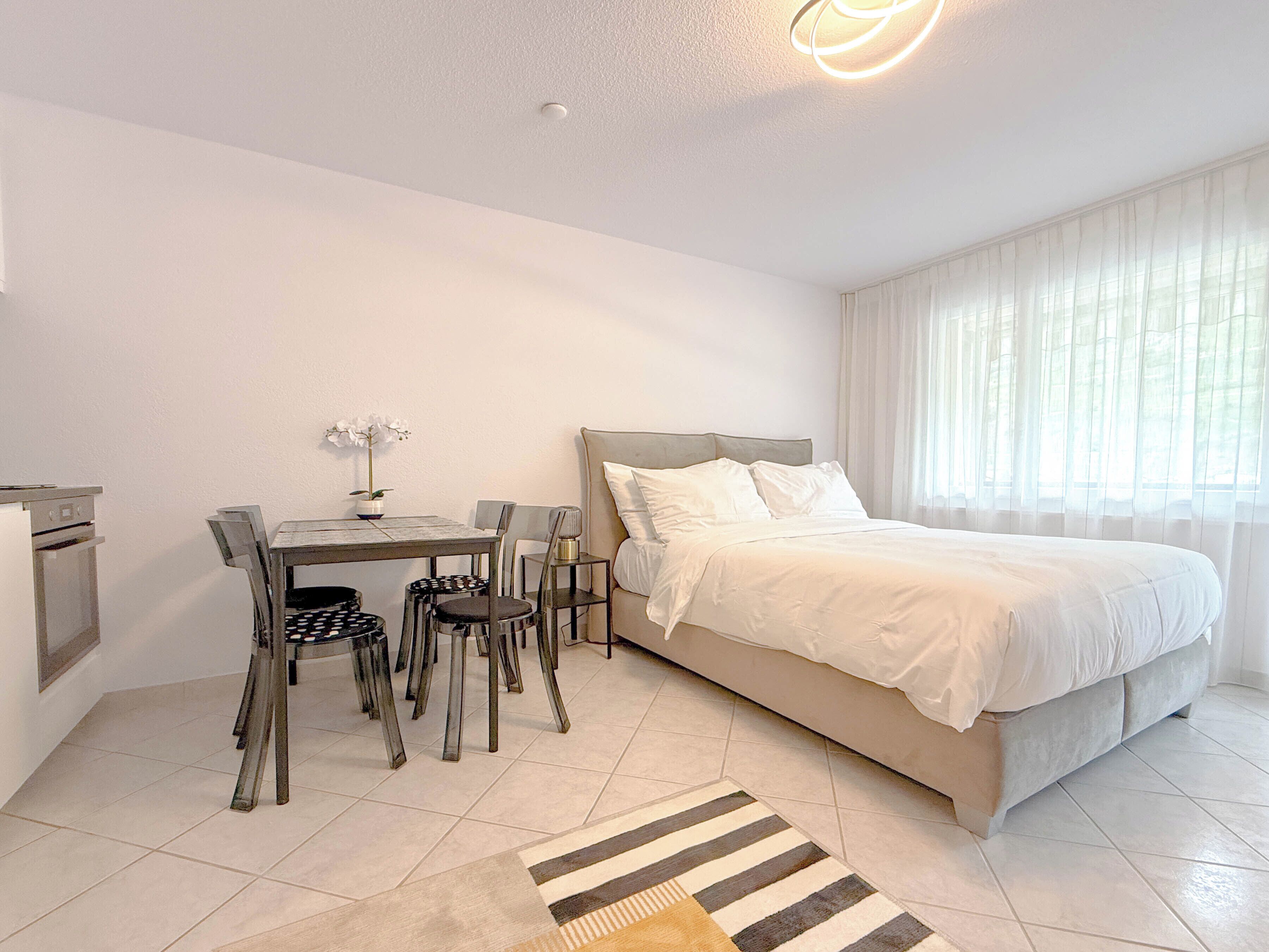 A bedroom with a bed, a desk, chairs, and a small table with a plant.