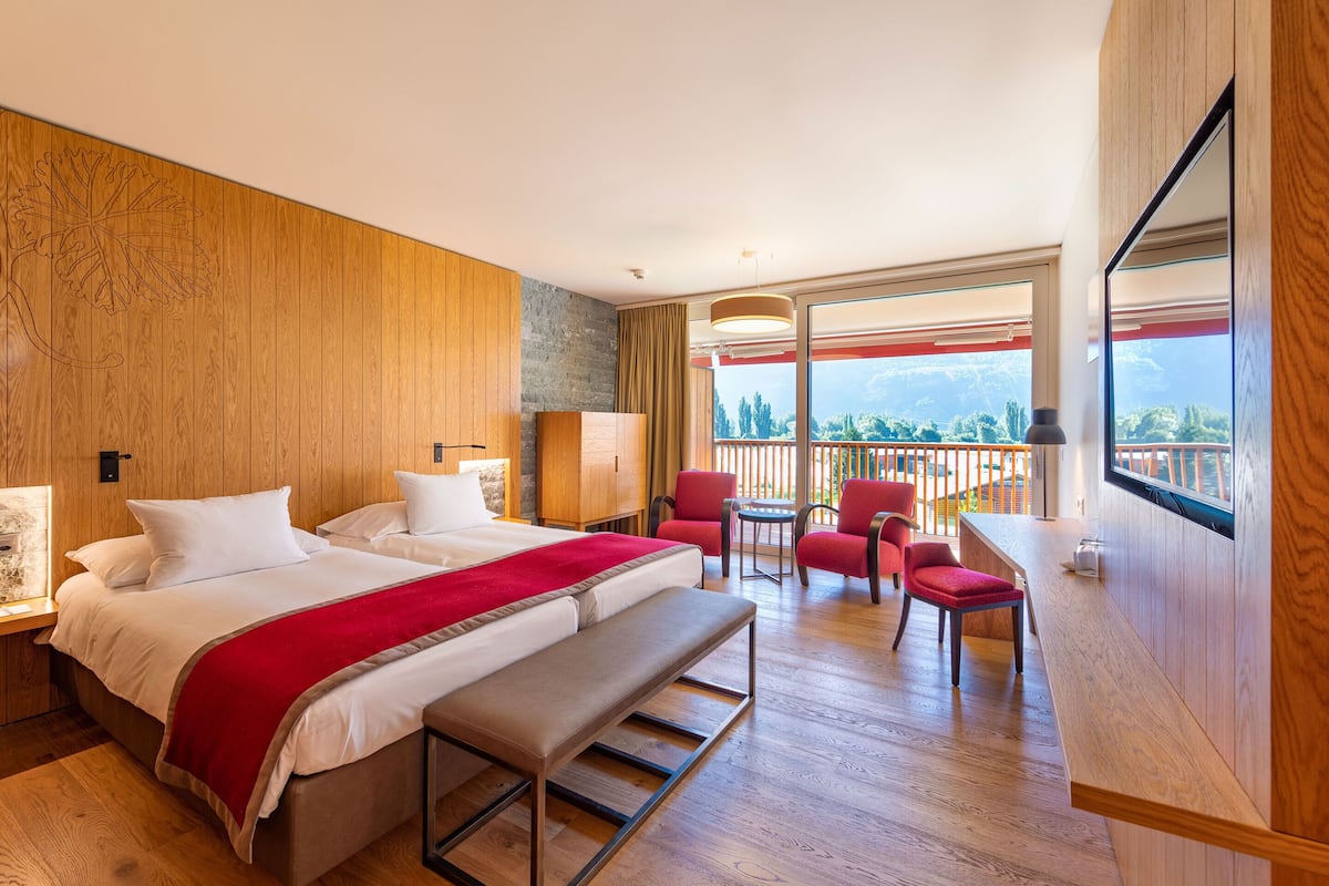 A hotel room with a large bed, a bench, a desk, and a balcony with red chairs.