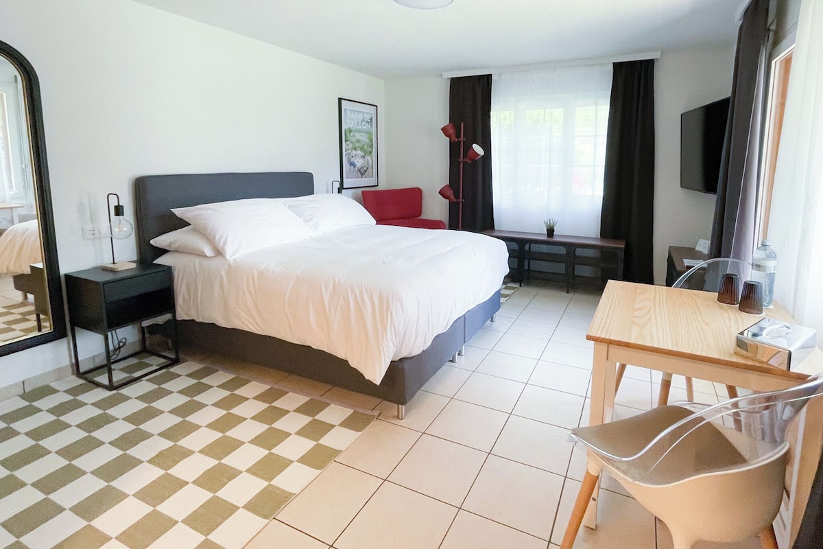 A hotel room with a bed, a desk, a chair, a mirror, a lamp, and a television.