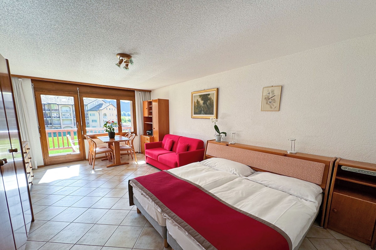 A hotel room with a large bed, a red sofa, a dining table with chairs, and a view of the outside through a sliding door.