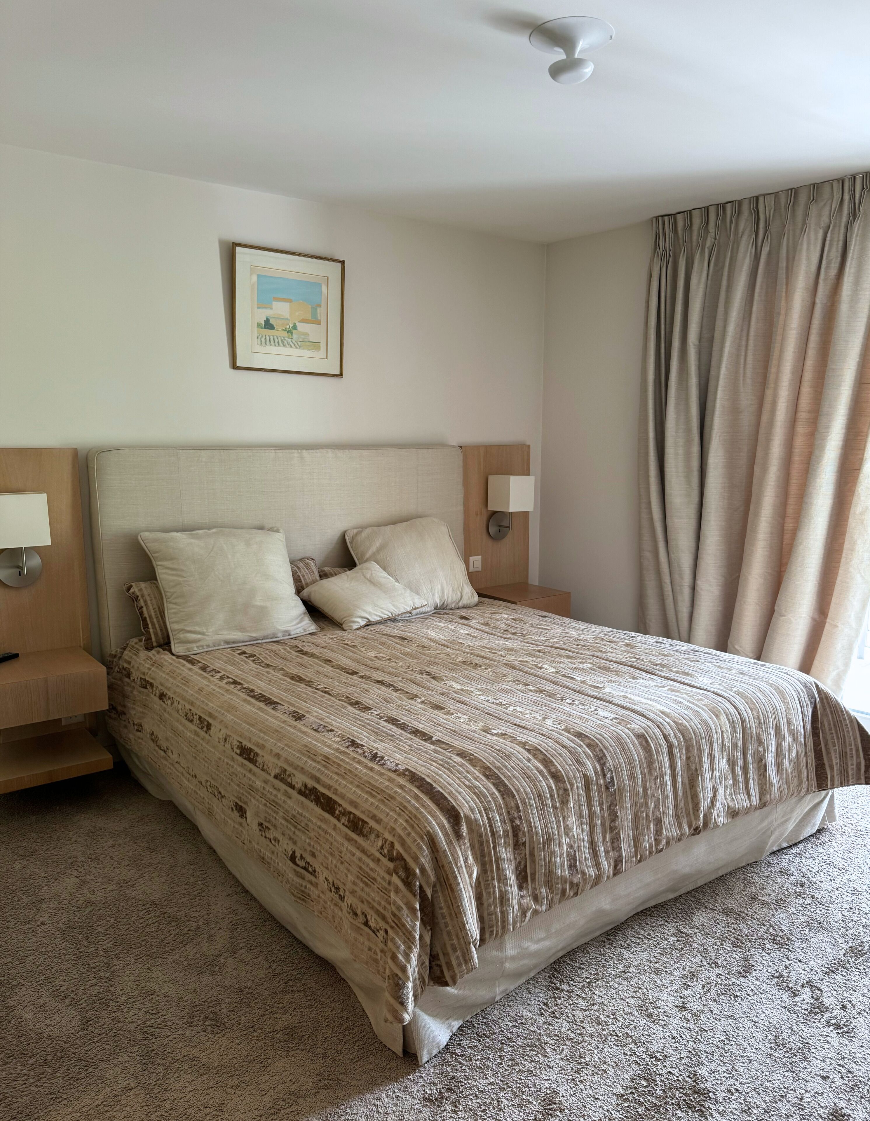 Una habitación de hotel con una cama, dos mesitas de noche, una lámpara, un cuadro en la pared y una ventana con cortinas.