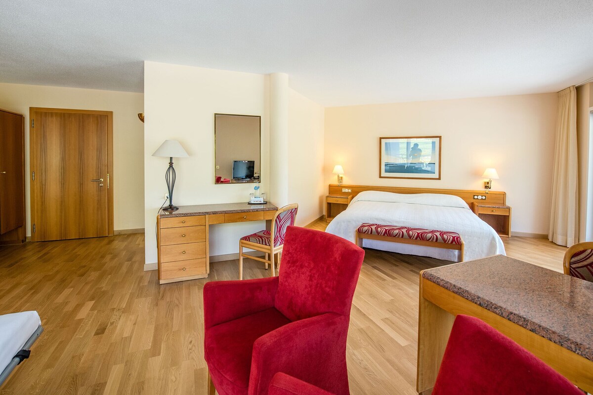 A hotel room with a bed, desk, chairs, a desk lamp, a bedside table, a TV, and a window with curtains.