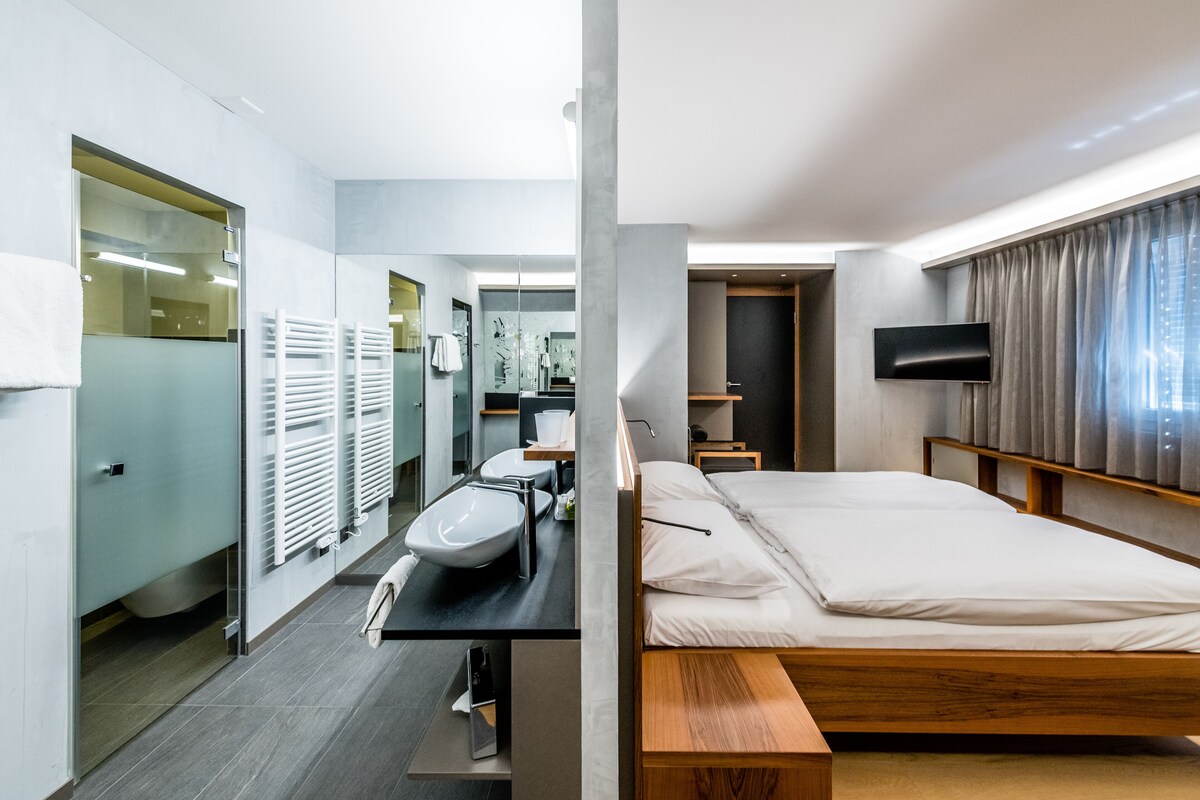 A modern hotel room with a wooden bed, a large mirror, a sink, and a flat-screen TV.