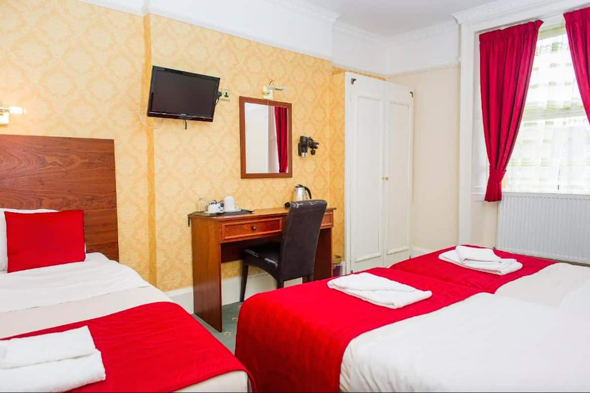 A hotel room with two beds, a desk, a chair, a television, and a window with red curtains.