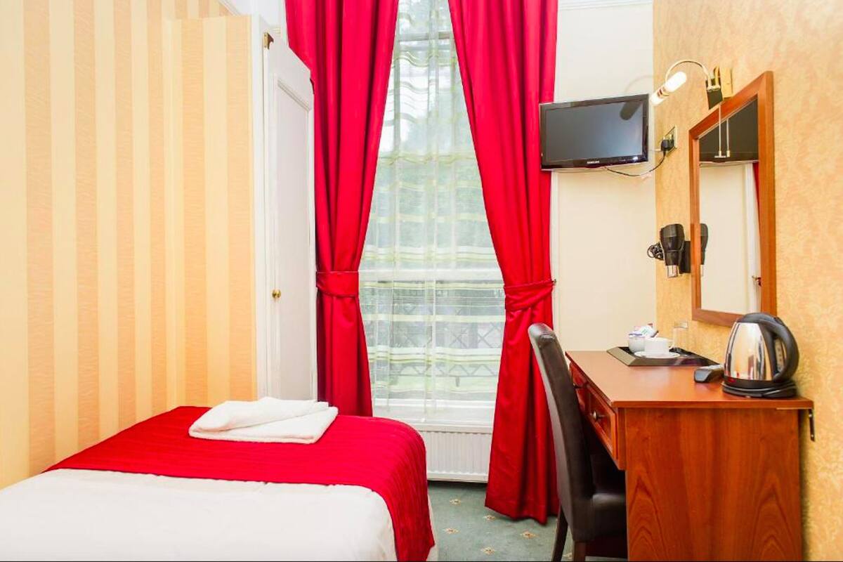 A hotel room with a bed, a desk with a kettle, a TV, and a window with red curtains.