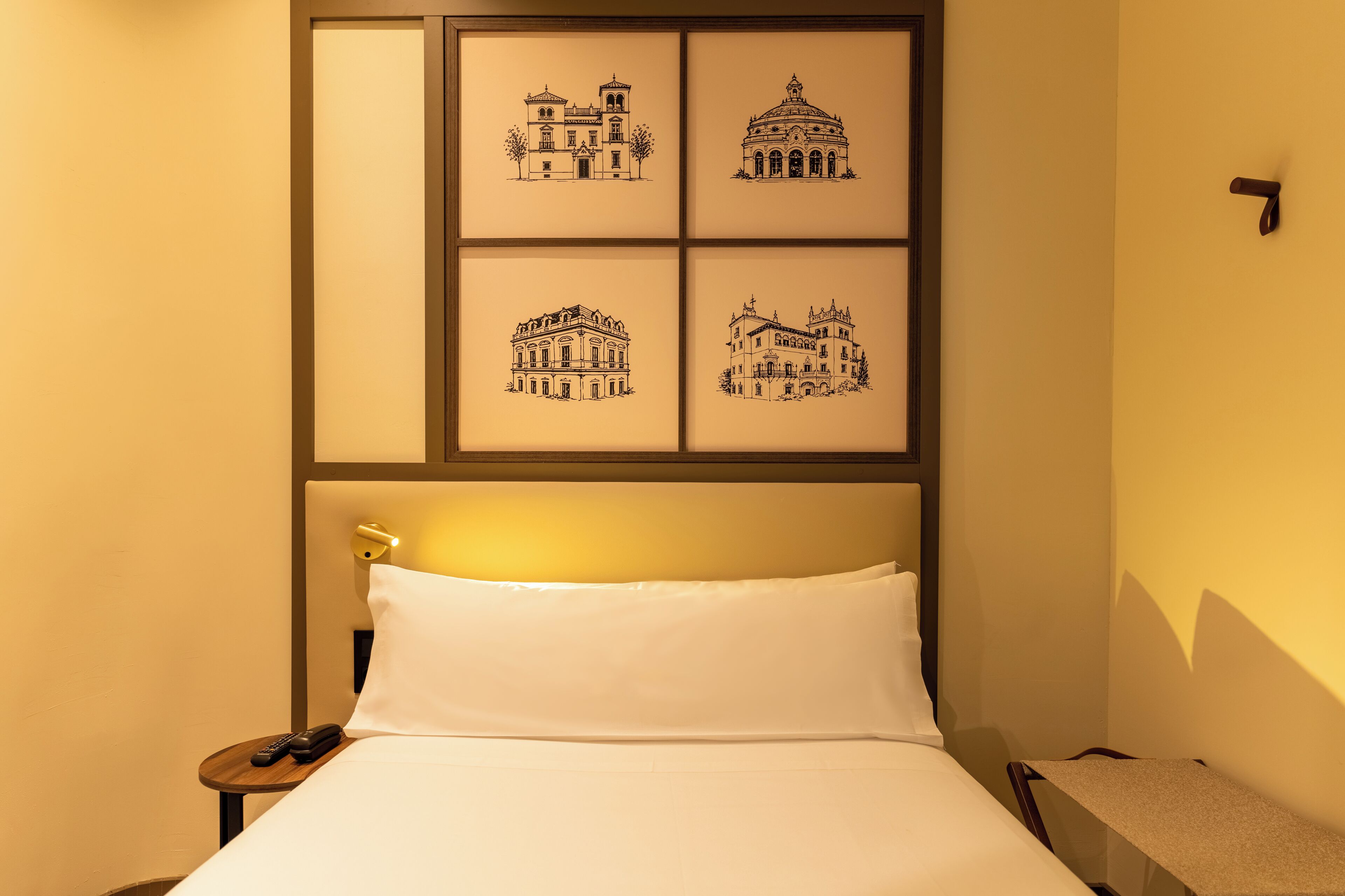 A hotel room with a bed, a nightstand, a telephone, and four framed black-and-white architectural drawings on the wall.