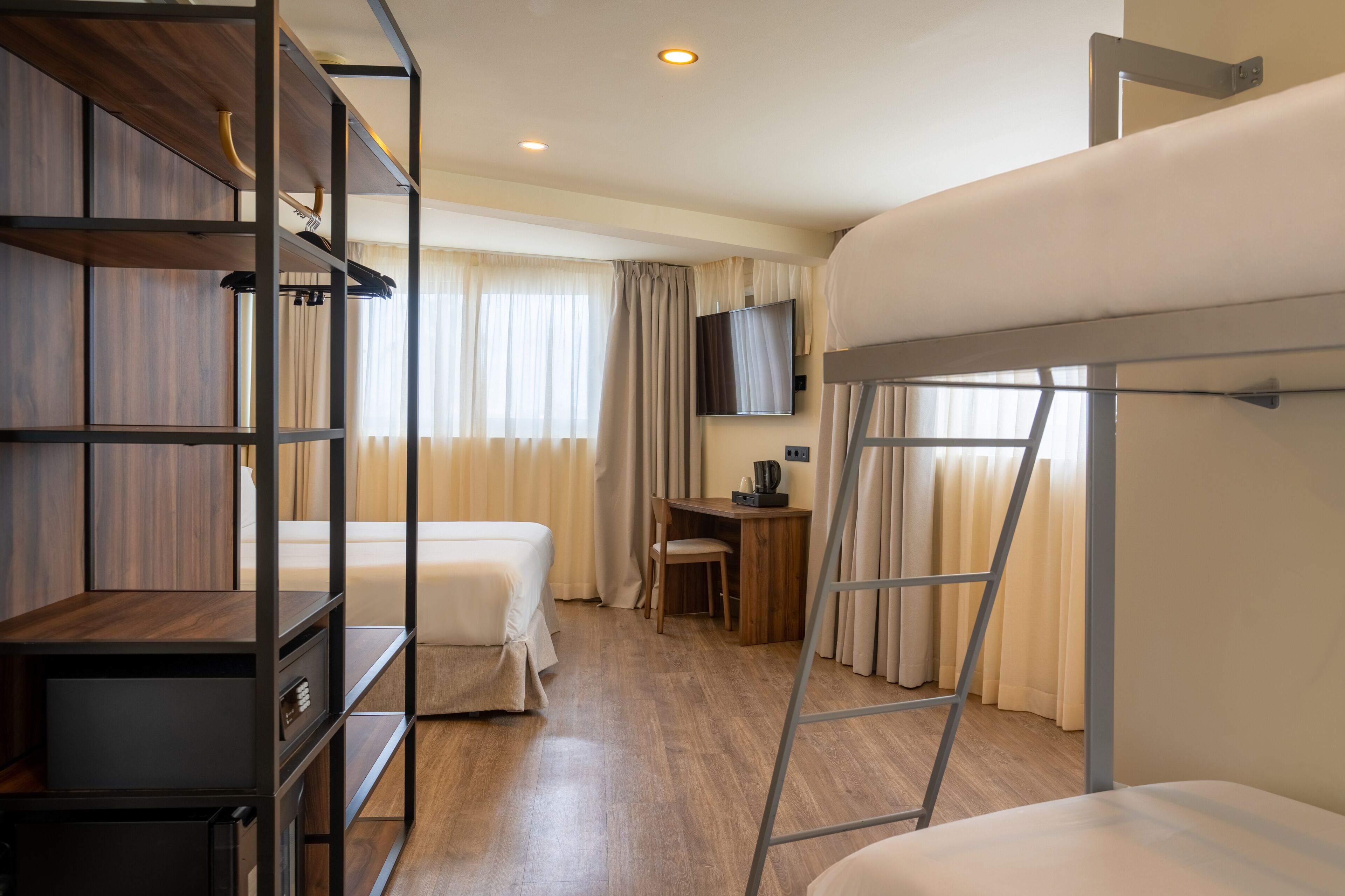 Une chambre d’hôtel avec des lits superposés, un bureau avec une chaise, un téléviseur fixé au mur et une armoire avec des étagères.