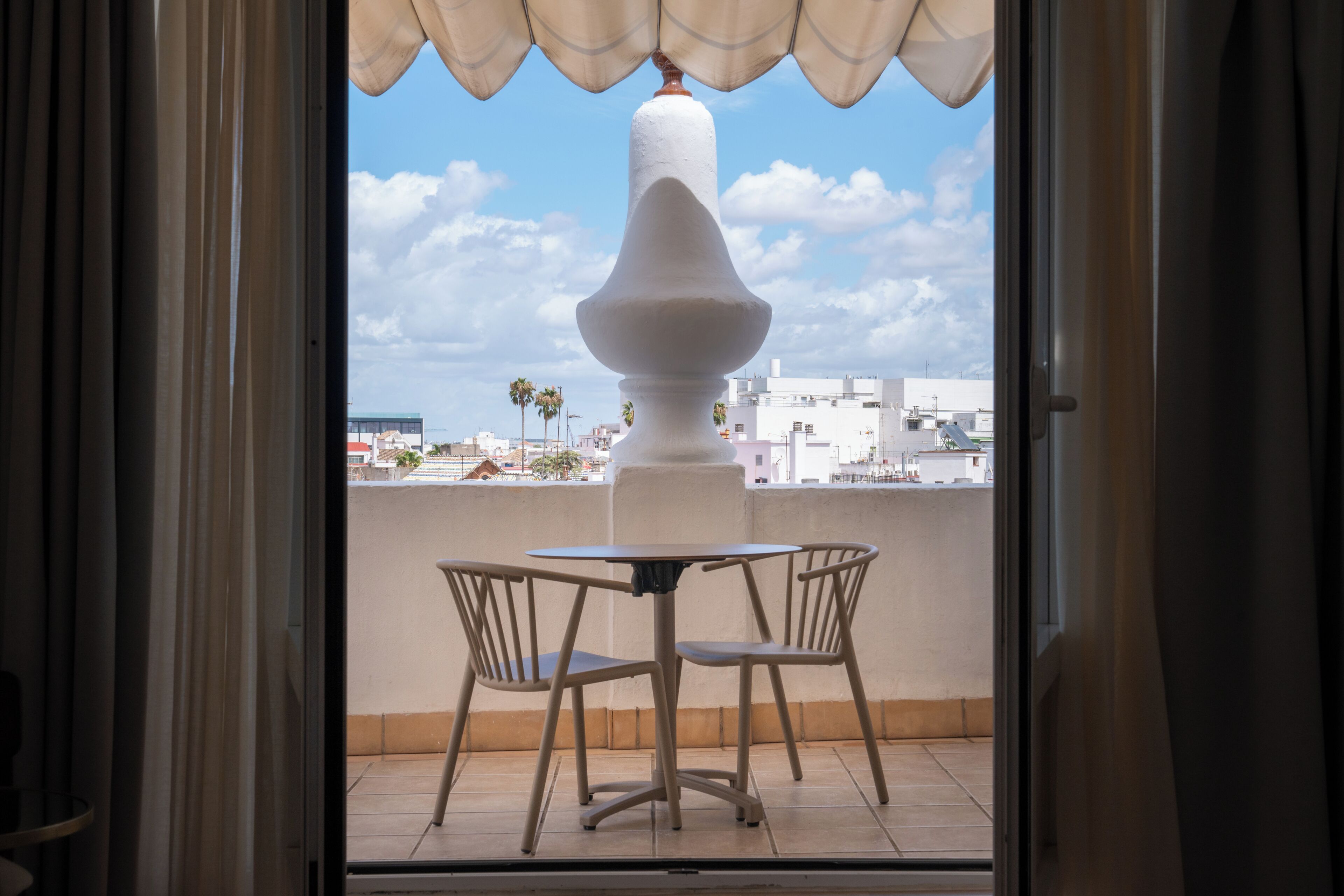 An open balcony with a table and chairs, a white column, and a view of buildings and palm trees.