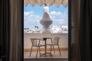 Un balcón abierto con una mesa y sillas, un pilar blanco y vistas a edificios y palmeras.