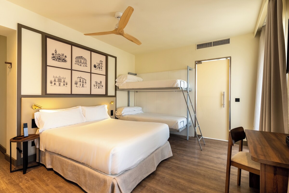 A hotel room with a large bed, a smaller bed on a bunk, a desk, a chair, a ceiling fan, and artwork on the wall.