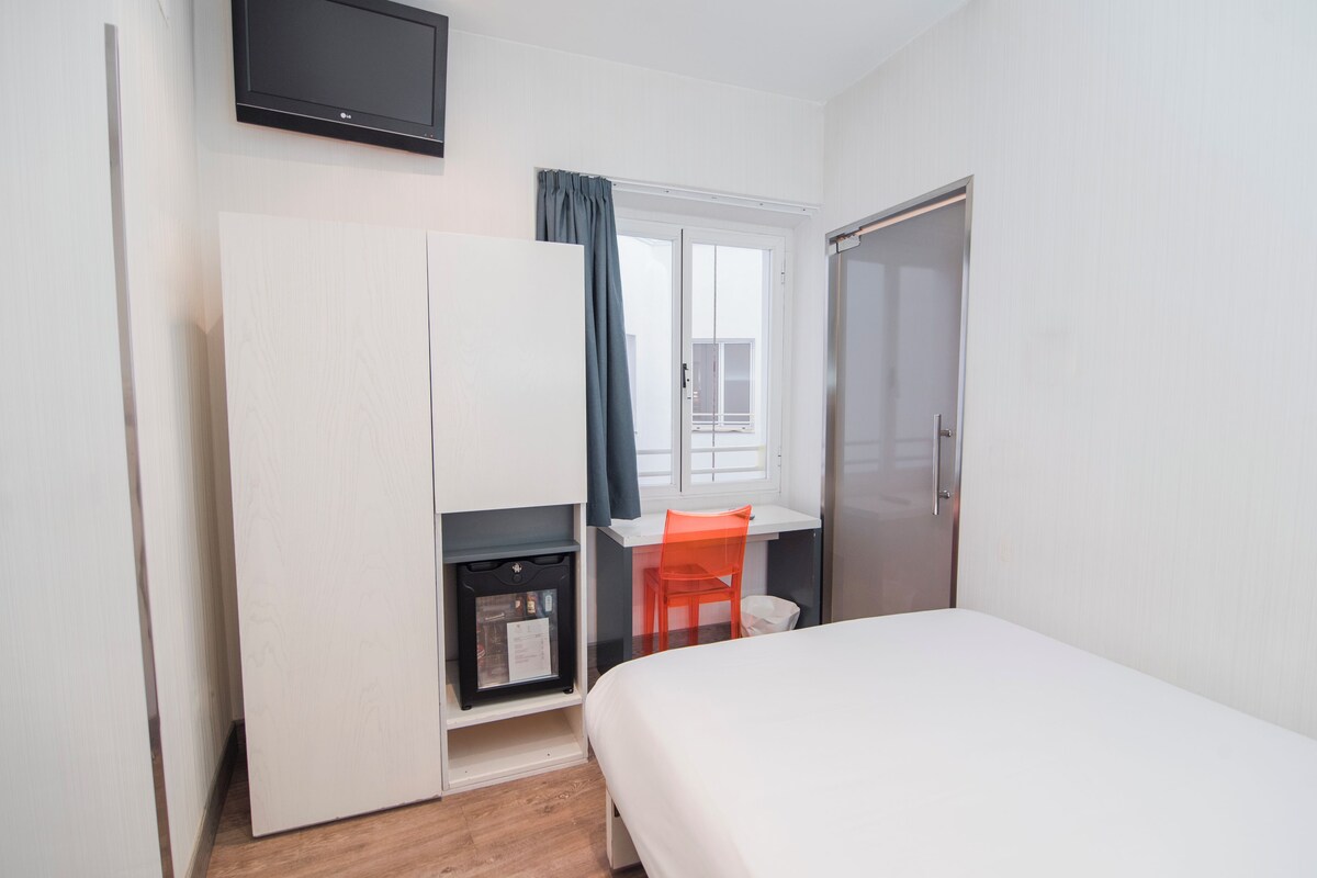 Un dormitorio con cama, escritorio y silla, armario, televisor y ventana con cortinas.