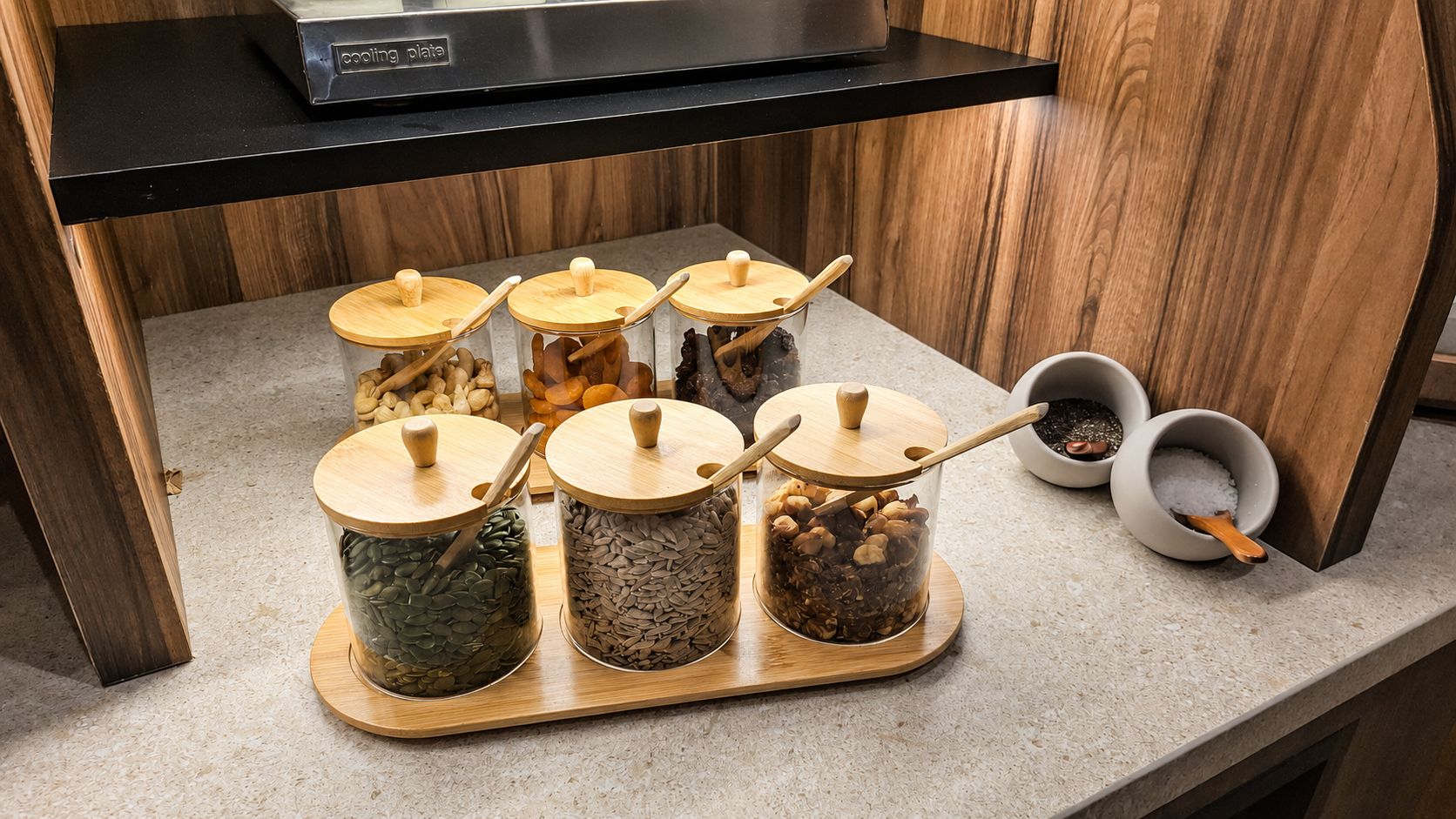 Een aanrecht met drie glazen potten gevuld met verschillende etenswaren, elk met een houten deksel en een houten lepel.