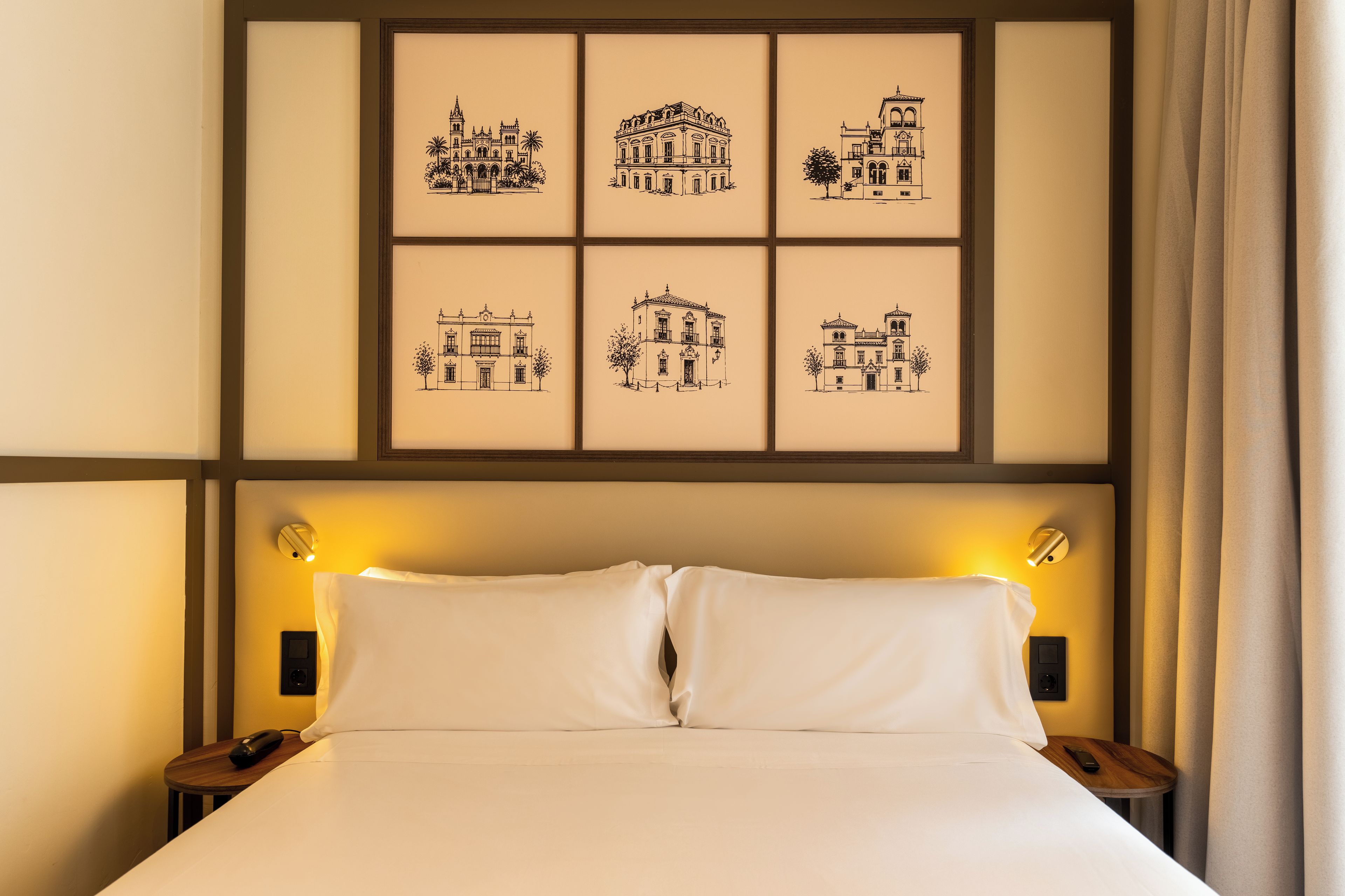 A hotel room with a bed, two bedside tables, and a wall with framed black-and-white architectural drawings.