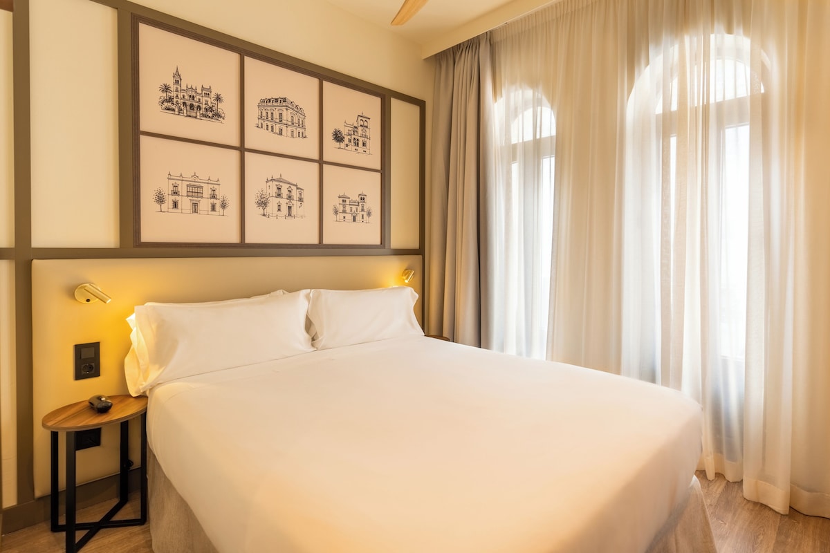 A hotel room with a large bed, a nightstand with a lamp, and a wall with framed black-and-white architectural drawings.