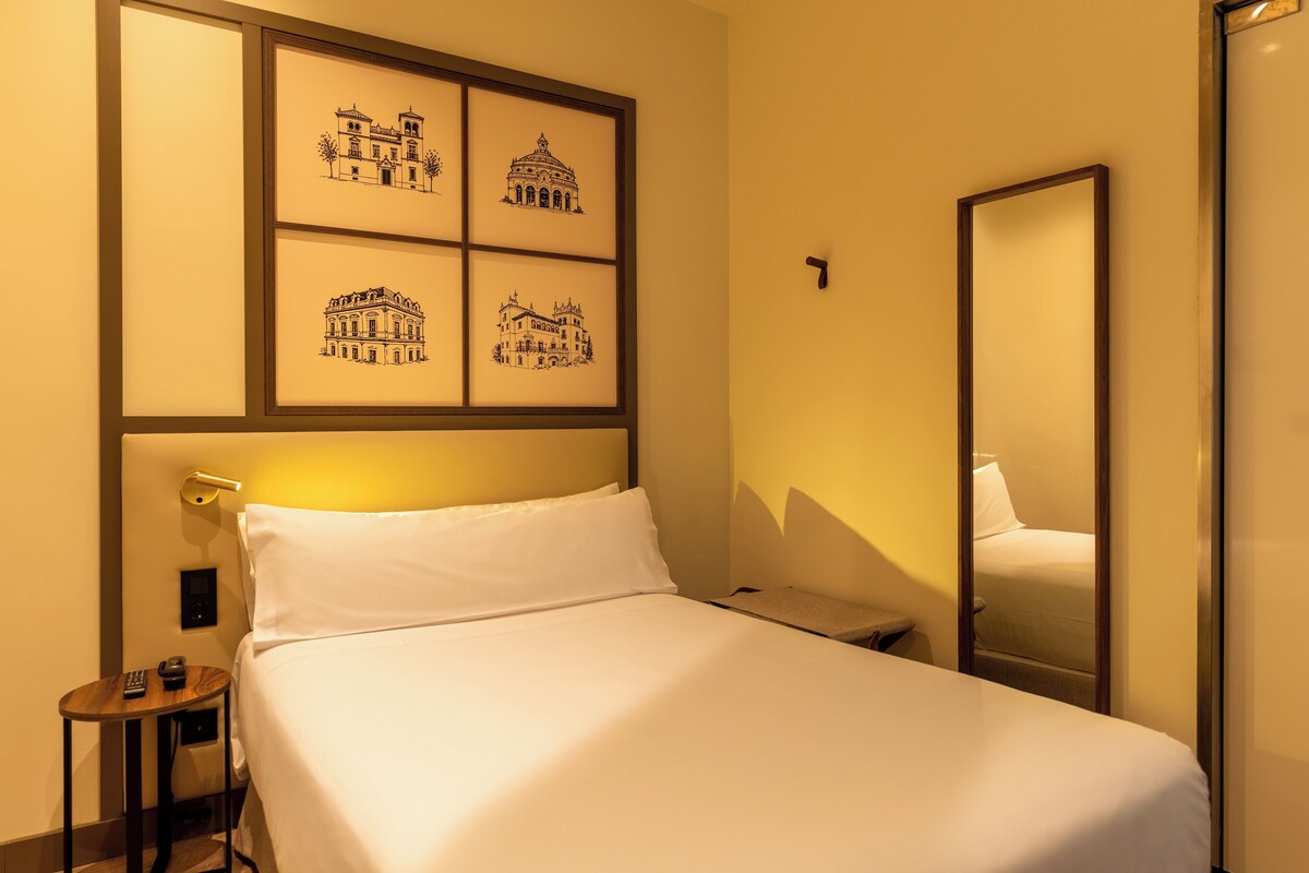 A hotel room with a bed, a bedside table, a small round table with a remote control, and a wall-mounted mirror.