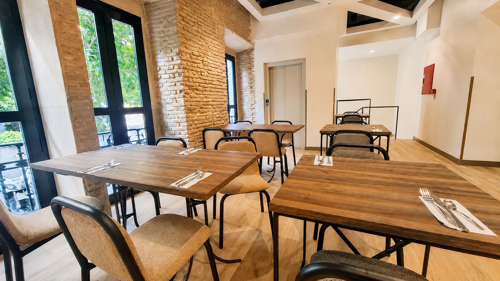 Een moderne eetruimte met houten tafels en stoelen, grote ramen en een accentmuur van baksteen.