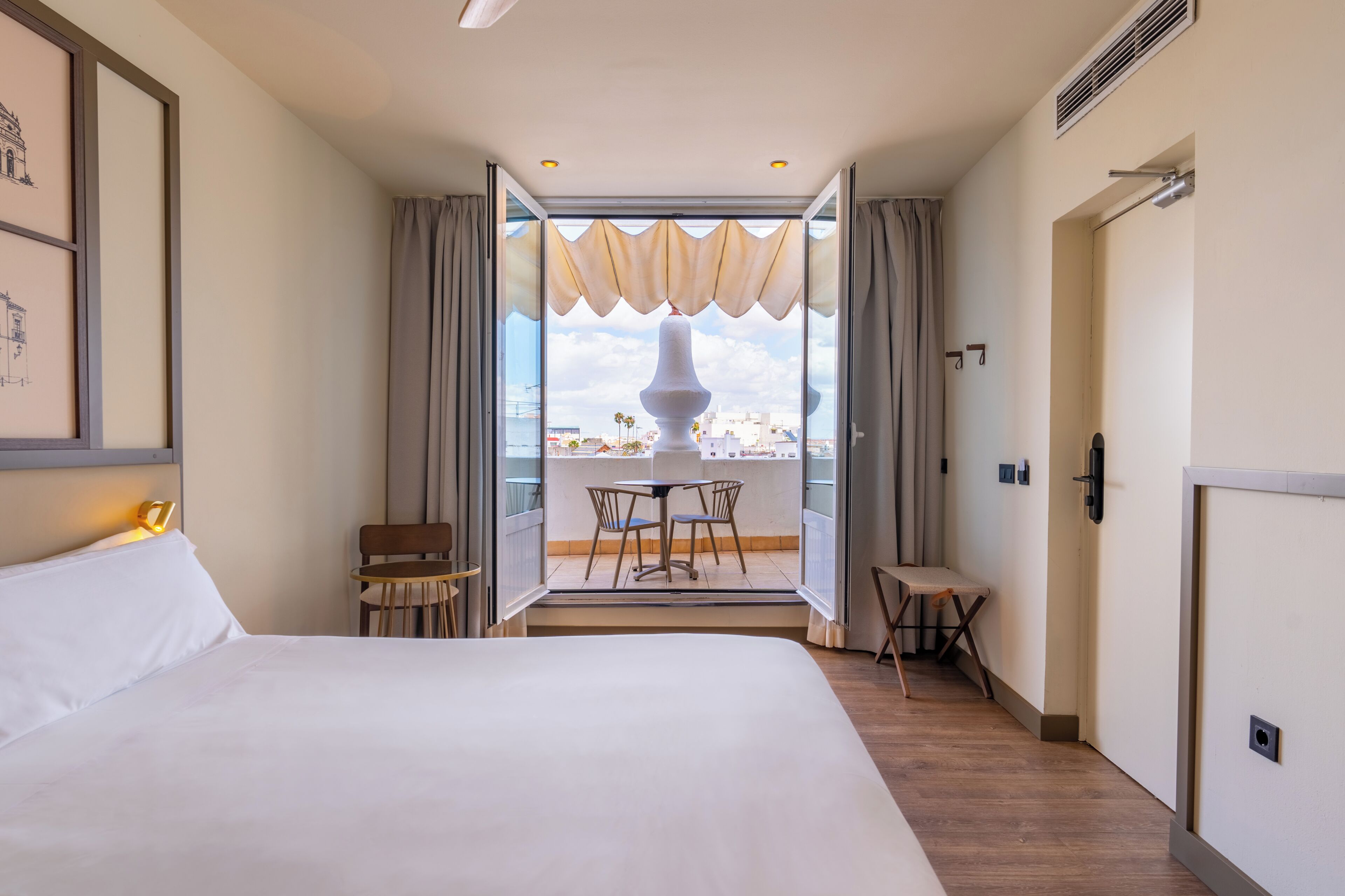 Habitación de hotel con una cama grande, un balcón con mesa y sillas, y vistas a la ciudad.