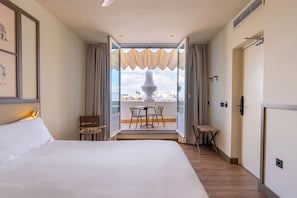 Habitación de hotel con una cama grande, un balcón con mesa y sillas, y vistas a la ciudad.
