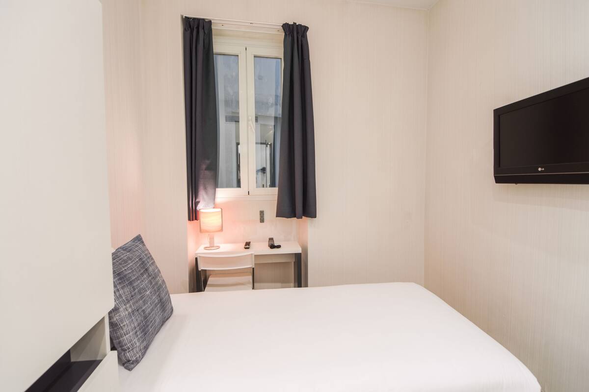 A hotel room with a bed, a desk, a lamp, a TV, and a window with curtains.