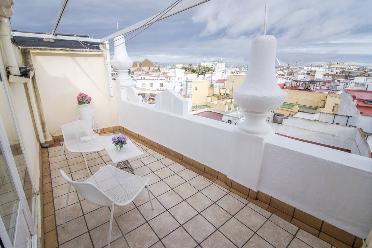 A balcony with two white chairs and a small table, overlooking a cityscape with buildings and a clear sky.