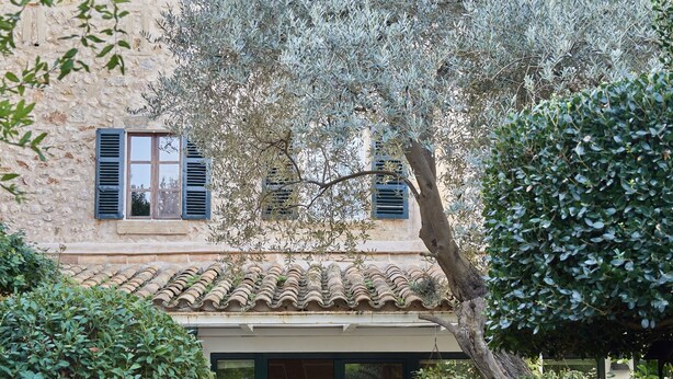 A traditional building with a tiled roof, a covered entrance, and a garden with various plants and a large tree. - Son Sant Jordi (Pollenca)