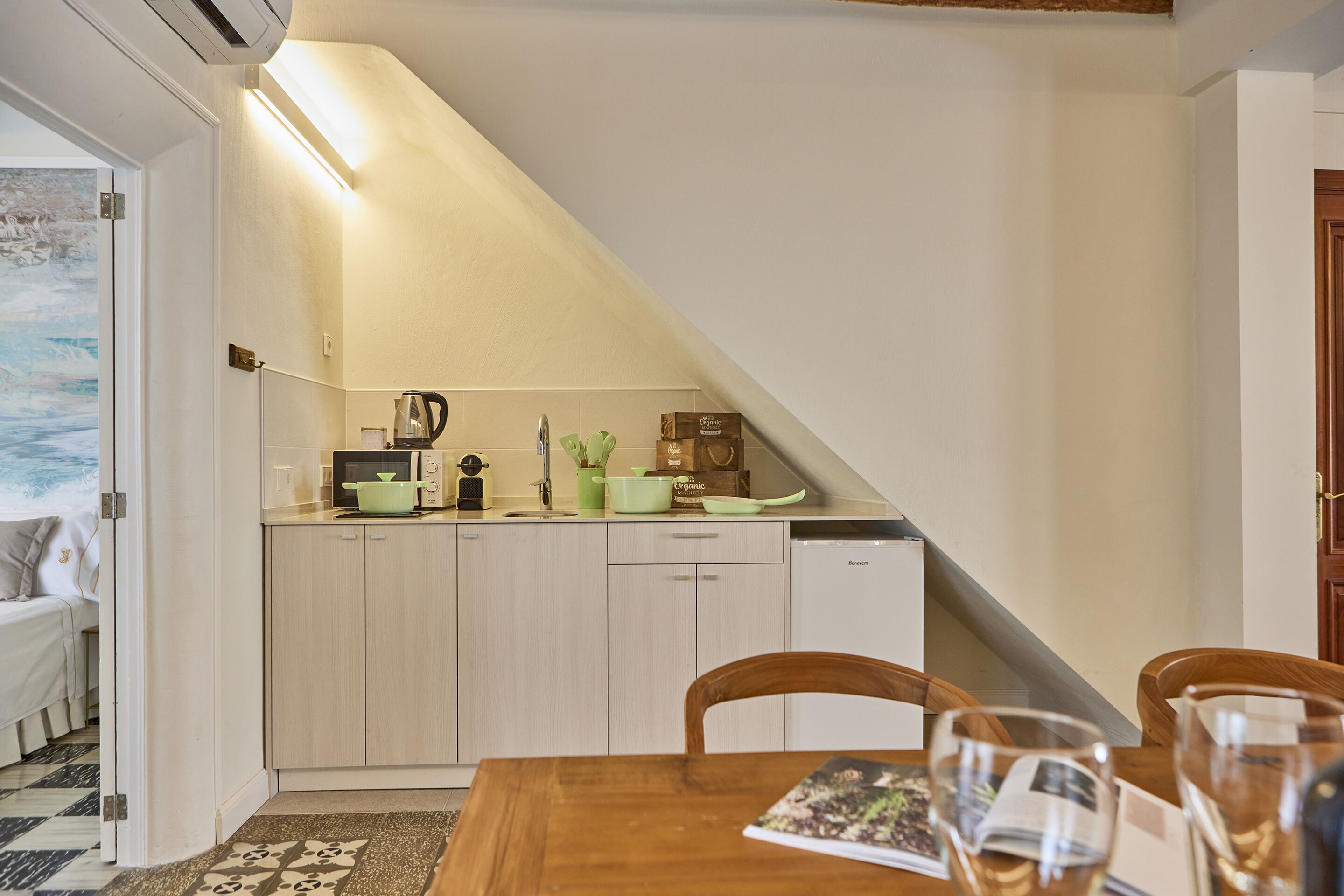 A compact kitchen with wooden cabinets, a dining table with chairs, and a view of a bedroom with a bed and a painting on the wall.