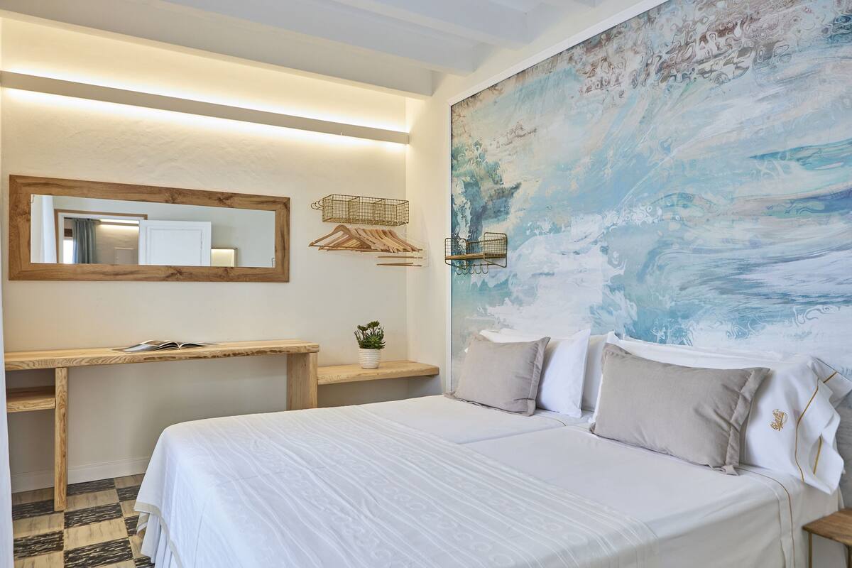 A bedroom with a bed, a wooden desk, a mirror, and a large abstract painting on the wall.