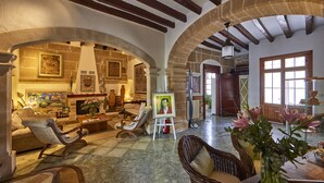 A spacious living area with high ceilings, exposed beams, and a fireplace. There are several seating options including a sofa, chairs, and a wicker armchair. Artwork is displayed on the walls, and a vase with flowers is on a table. - Son Sant Jordi (Pollenca)