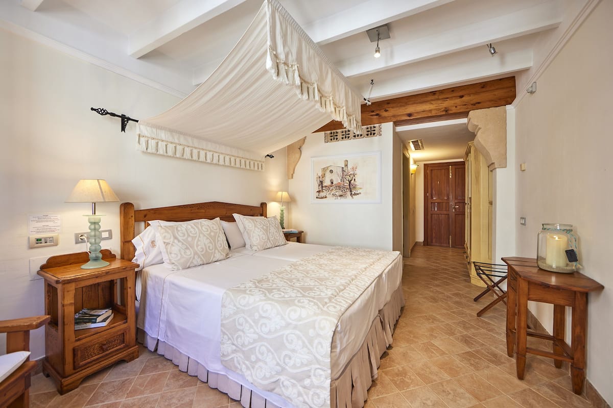 A bedroom with a large bed, wooden headboard, bedside tables, a hanging canopy, and a view of the hallway.