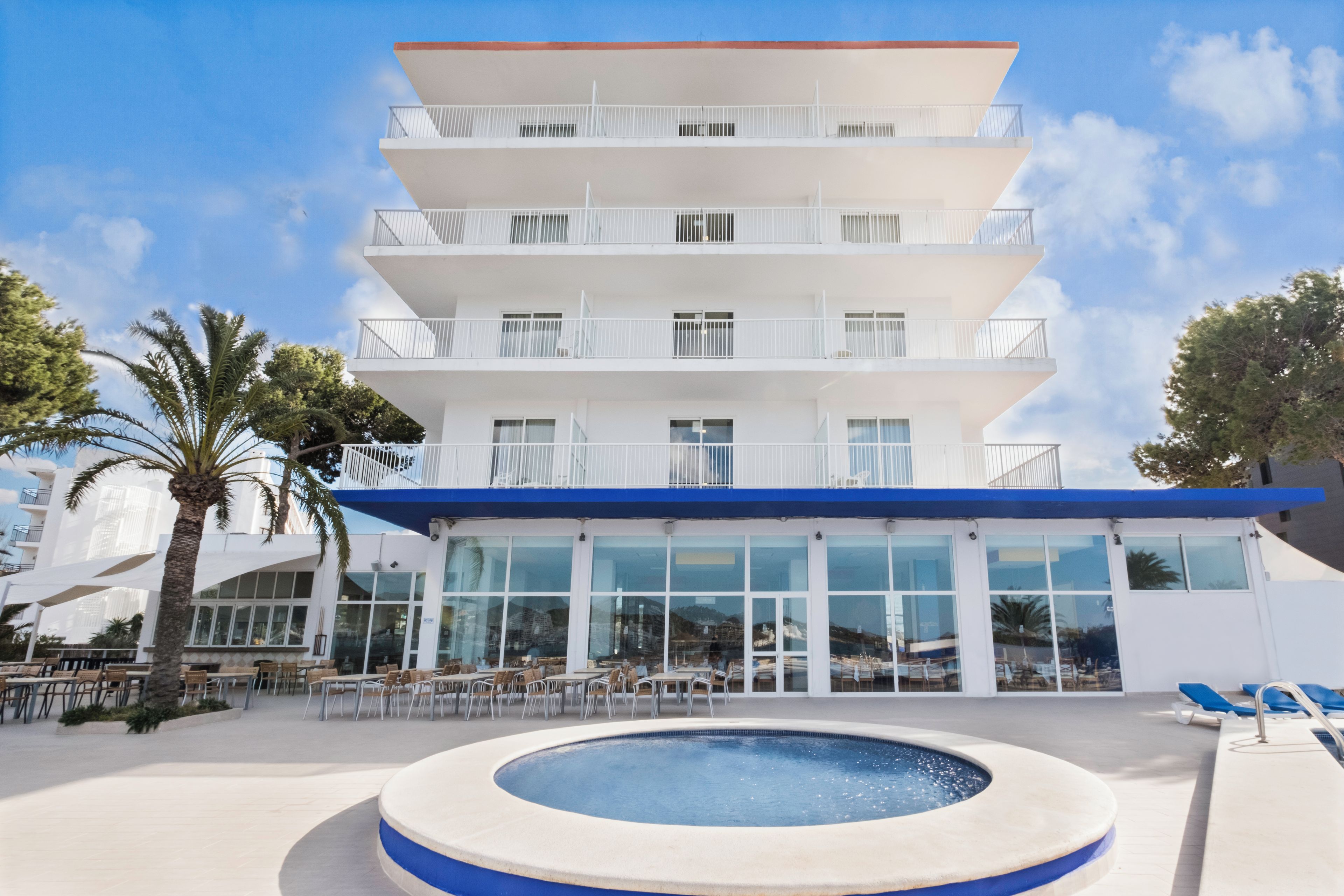 Un hotel de varios pisos con piscina, mobiliario exterior y cielo despejado.