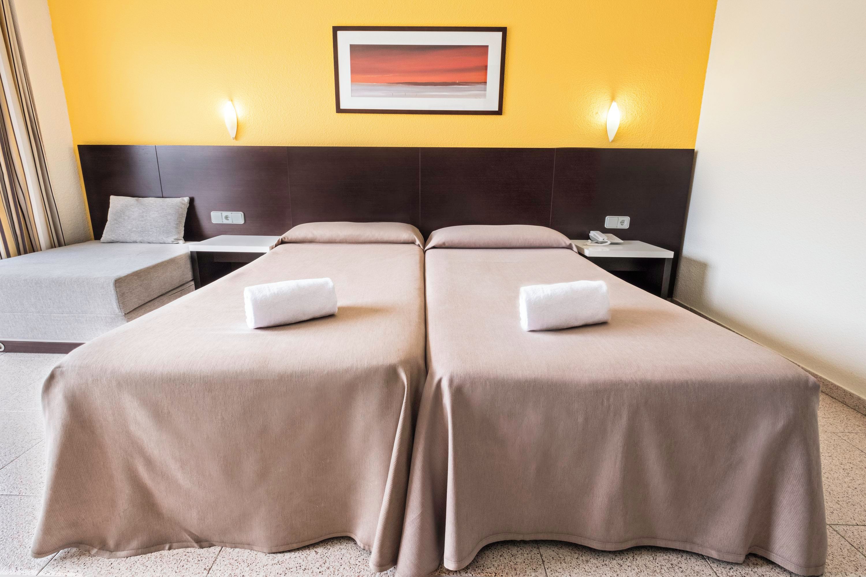 A hotel room with two beds, a nightstand, a bedside table, a framed picture on the wall, and a light switch.