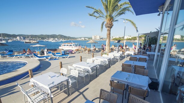 Un área de comedor al aire libre con mesas y sillas, una piscina y vistas a barcos y al agua.