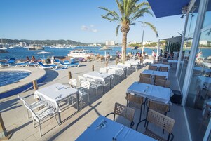 Un área de comedor al aire libre con mesas y sillas, una piscina y vistas a barcos y al agua.