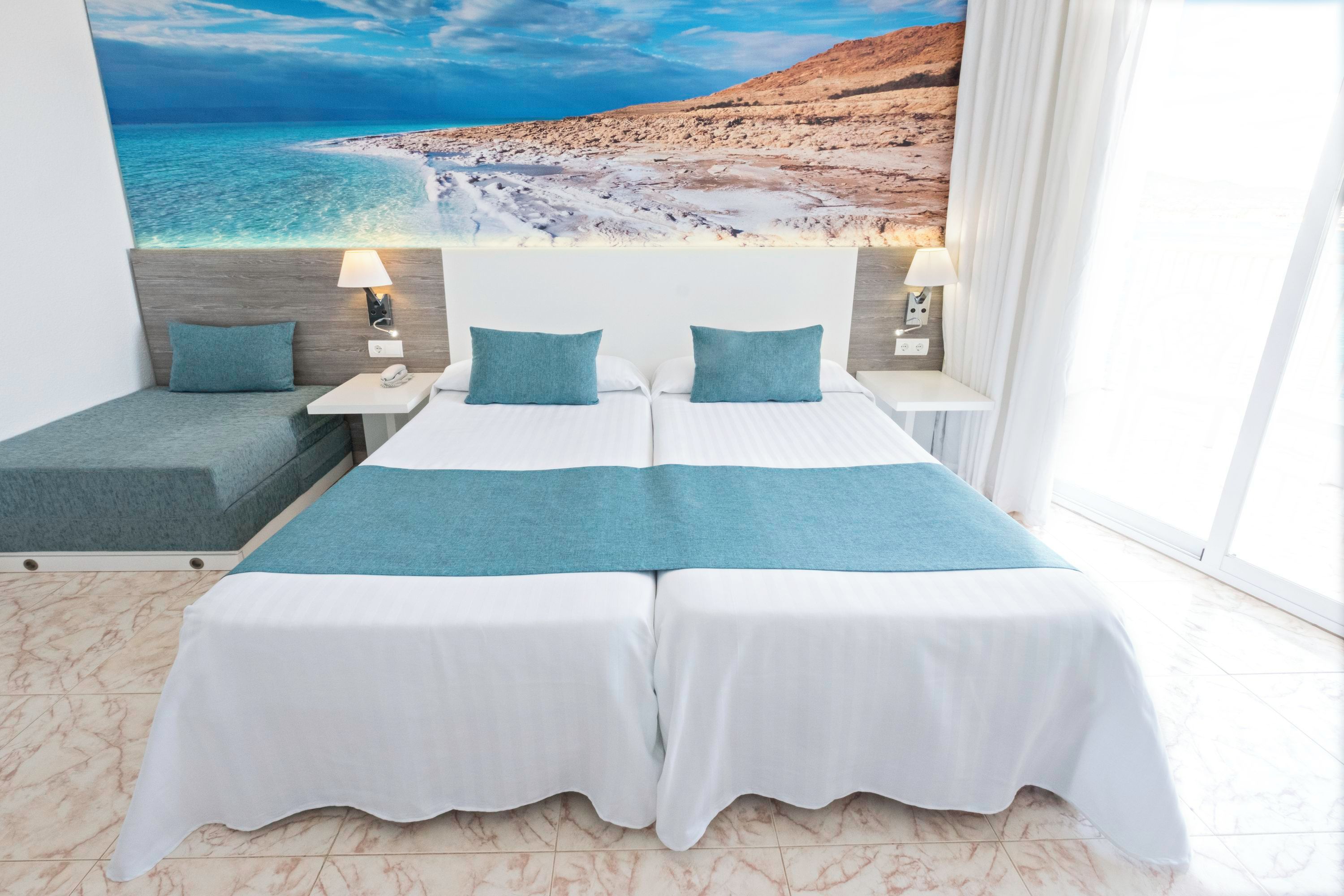 A double bed with a white and teal bedspread, two matching pillows, a bedside table with a lamp, and a wall mural of a beach scene.