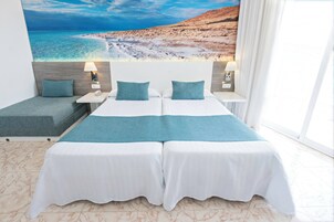 Una cama doble con una colcha blanca y azul turquesa, dos almohadas a juego, una mesita de noche con una lámpara y un mural en la pared con una escena de playa.