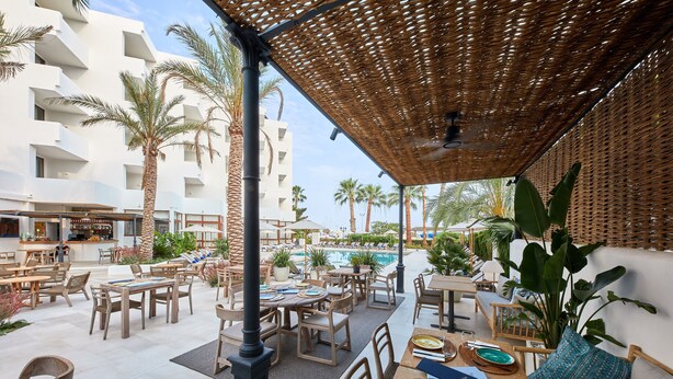 Un área de comedor al aire libre con mesas y sillas, una piscina y palmeras.