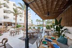 Un área de comedor al aire libre con mesas y sillas, una piscina y palmeras.